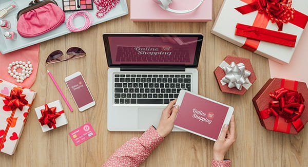 An top view of a desk with a person holding tablet. Also seen are a laptop and a smartphone along with various gifts.