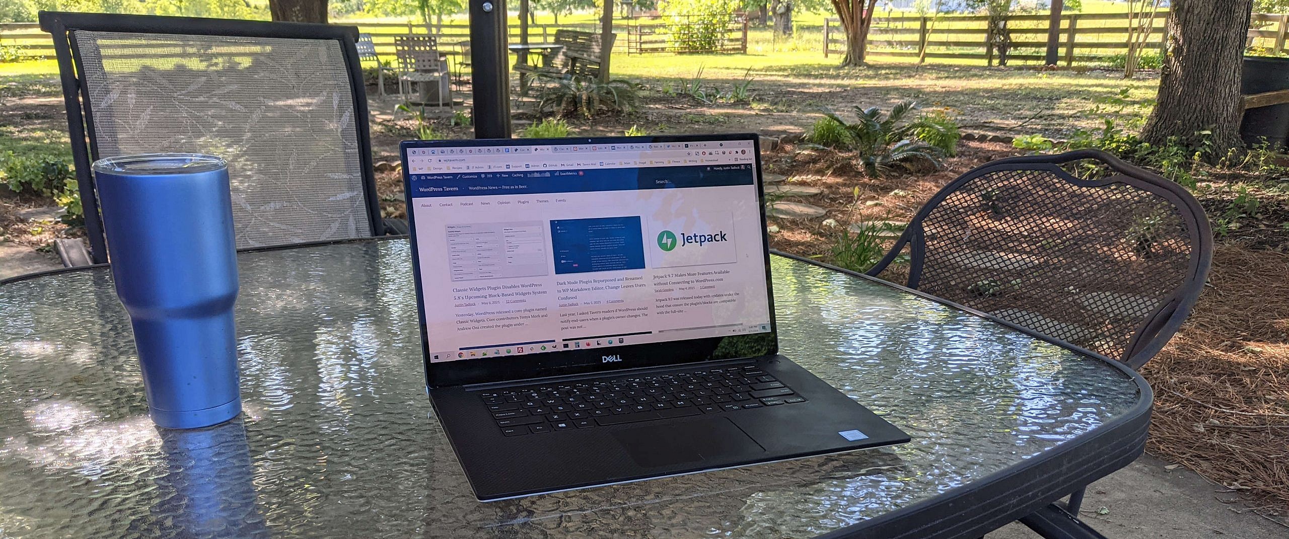 A laptop sitting on top of a glass table on a patio. Garden, swing, and trees in the background.